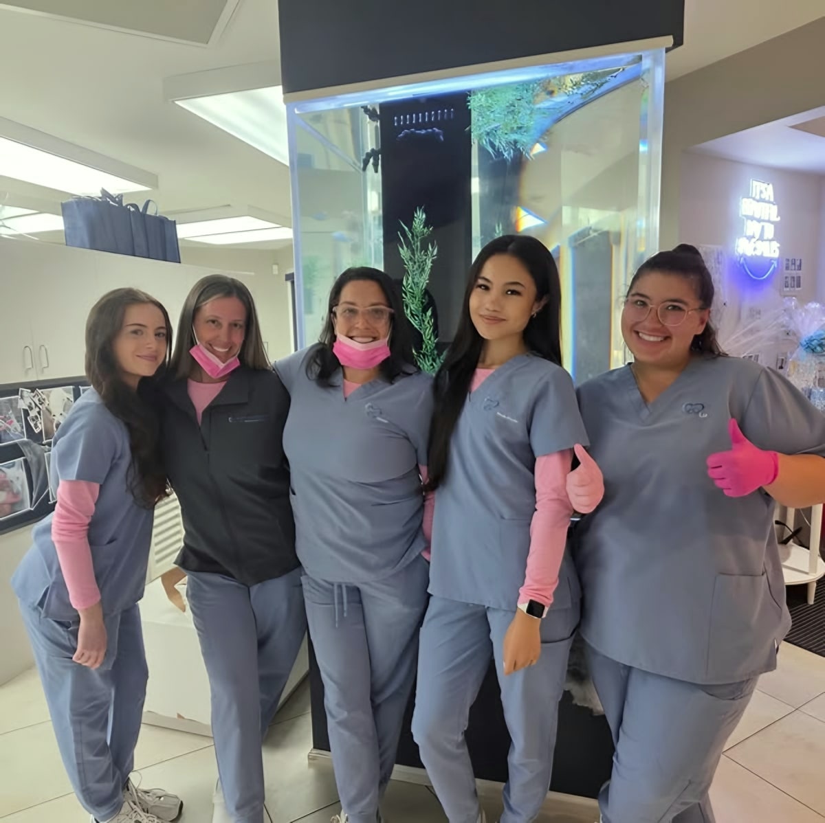 Five staff in scrubs posing by office fish tank, thumbs up
